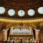 Orchestra rehearsing, in street clothes, on a concert hall stage with a pipe organ in the background