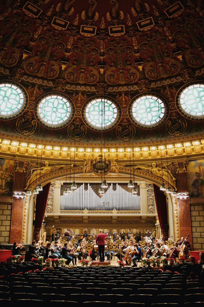 Orchestra rehearsing, in street clothes, on a concert hall stage with a pipe organ in the background