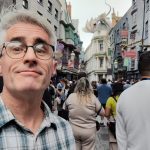 Quin Swallow on the Diagon Alley Street, with Gringotts Bank in the background (with the sculpted dragon on the roof), and a crowd of tourists behind him. Quin, with gray medium length hair parted and swept to the side, is wearing dark roundish glasses, and a green and tan plaid shirt. It is daytime, and the weather is clear.