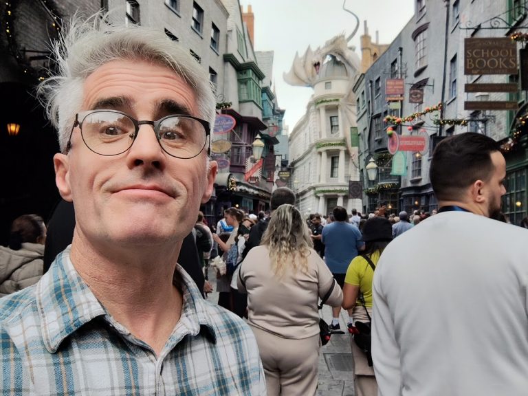 Quin Swallow on the Diagon Alley Street, with Gringotts Bank in the background (with the sculpted dragon on the roof), and a crowd of tourists behind him. Quin, with gray medium length hair parted and swept to the side, is wearing dark roundish glasses, and a green and tan plaid shirt. It is daytime, and the weather is clear.